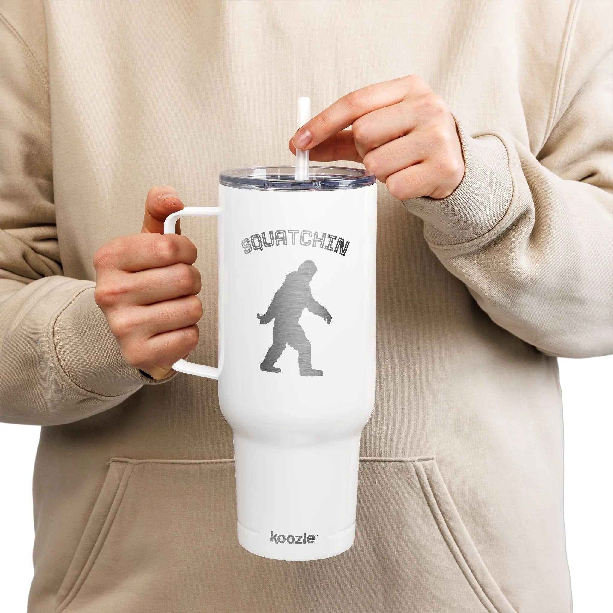 Person holding a white mug with 'SQUATCHIN' and a Bigfoot silhouette, branded 'koozie', against a beige background.