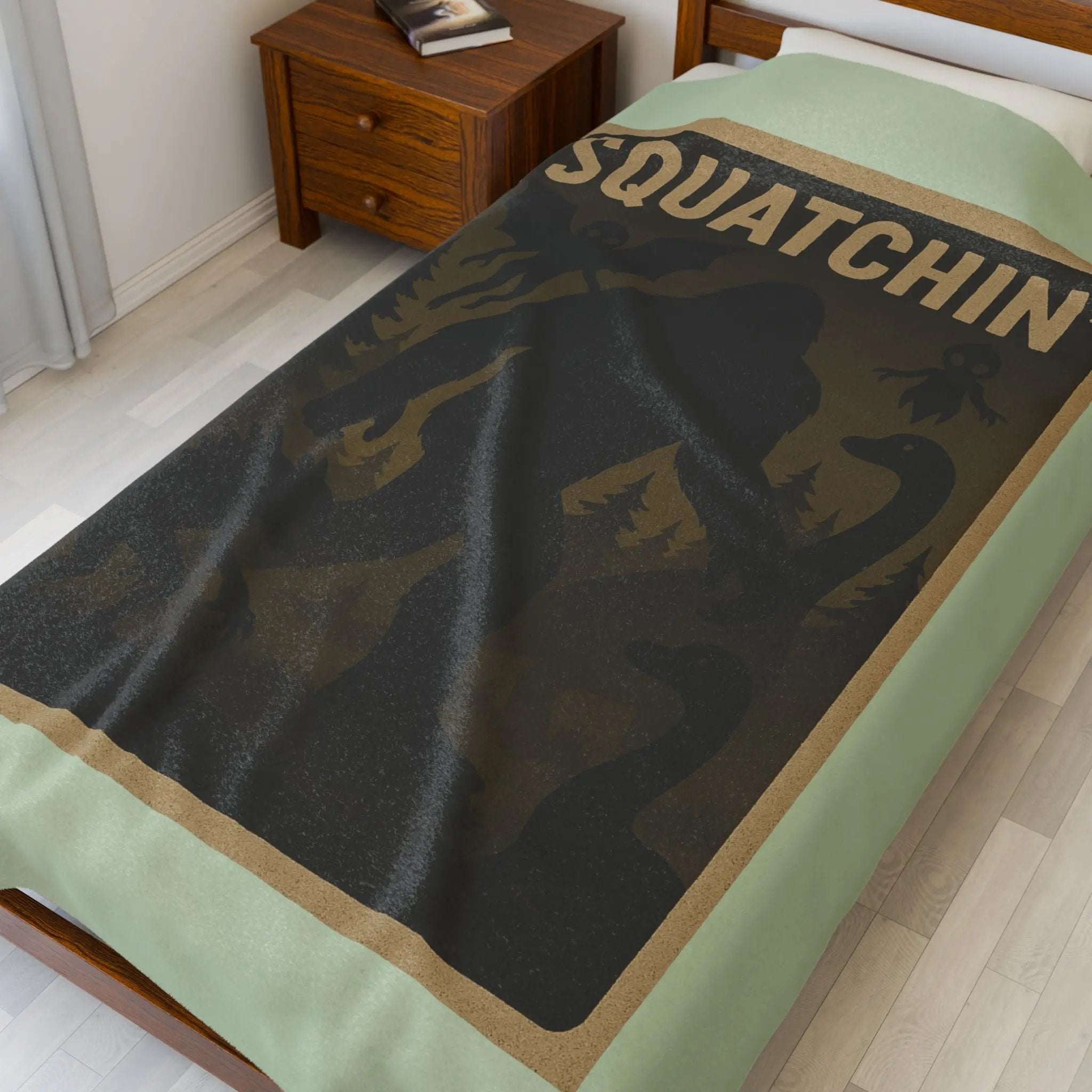 Light green fleece blanket with 'Squatchin' text across the top with silhouettes of bigfoot, loch Ness, werewolf, mothman, and an alien on a bed in a bedroom setting.