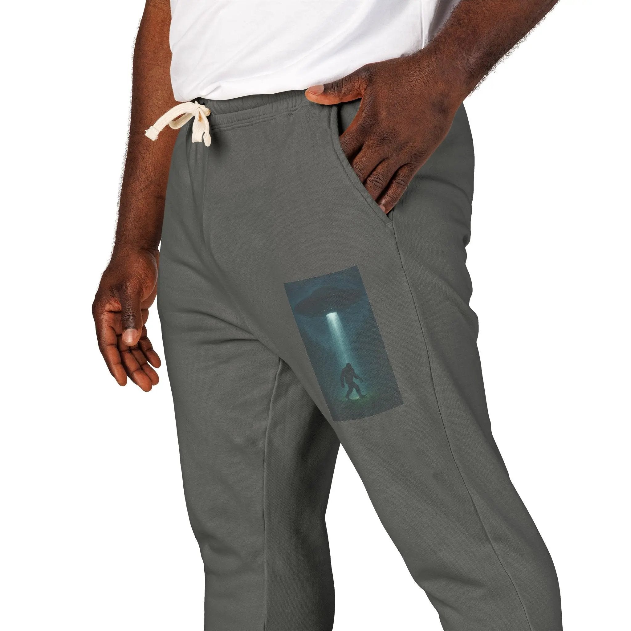 Gray sweatpants with a small graphic of a UFO with a beam of light on a bigfoot silhouette on the left leg worn by a person.