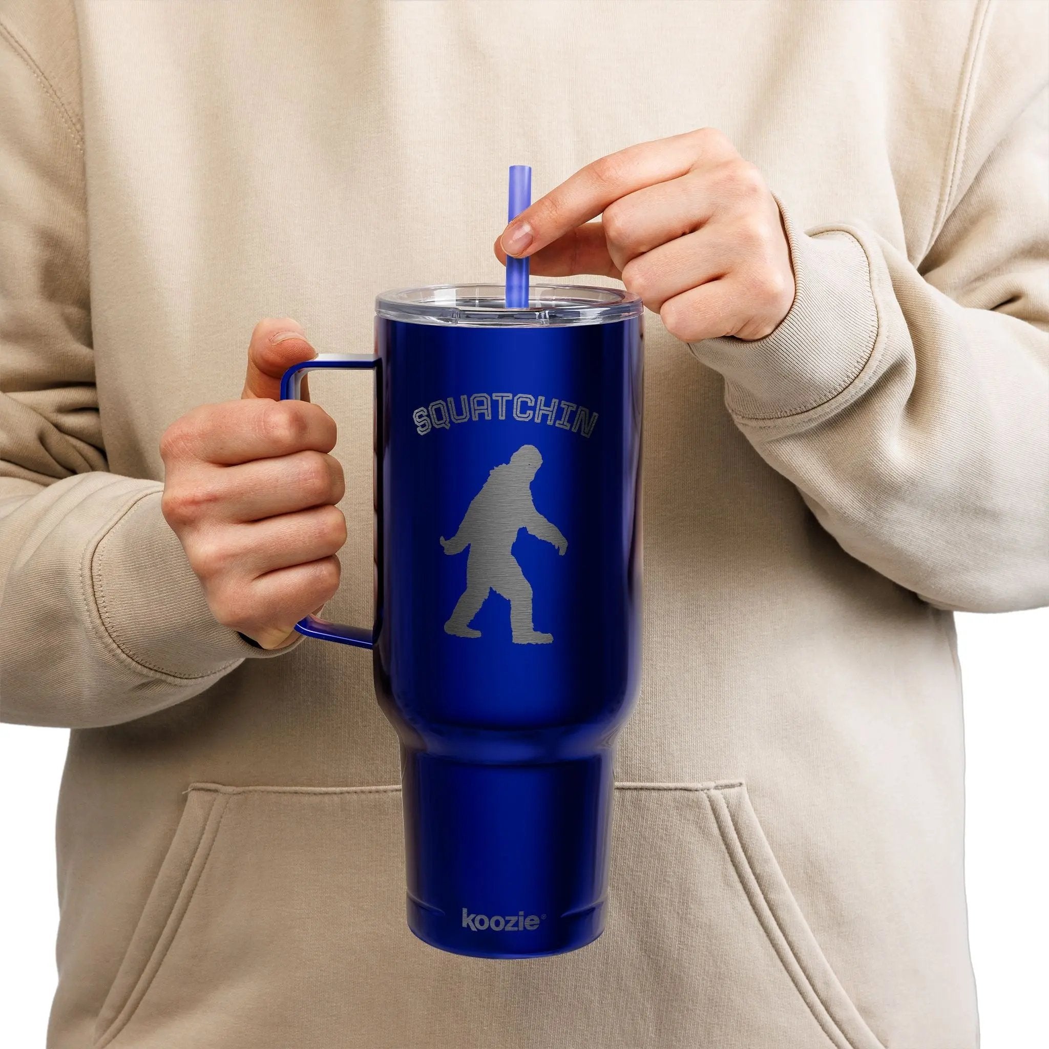 Person holding a blue mug with 'SQUATCHIN' and a silhouette of a bigfoot, against a beige background.