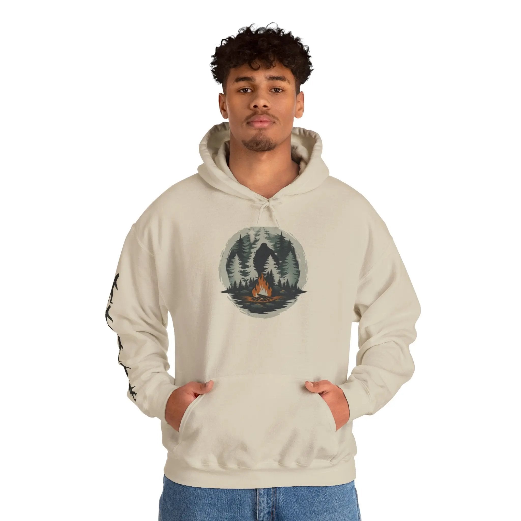 Person wearing a beige hoodie with a forest and campfire design and black bigfoot silhouettes on the right sleeve on a white background on a white background