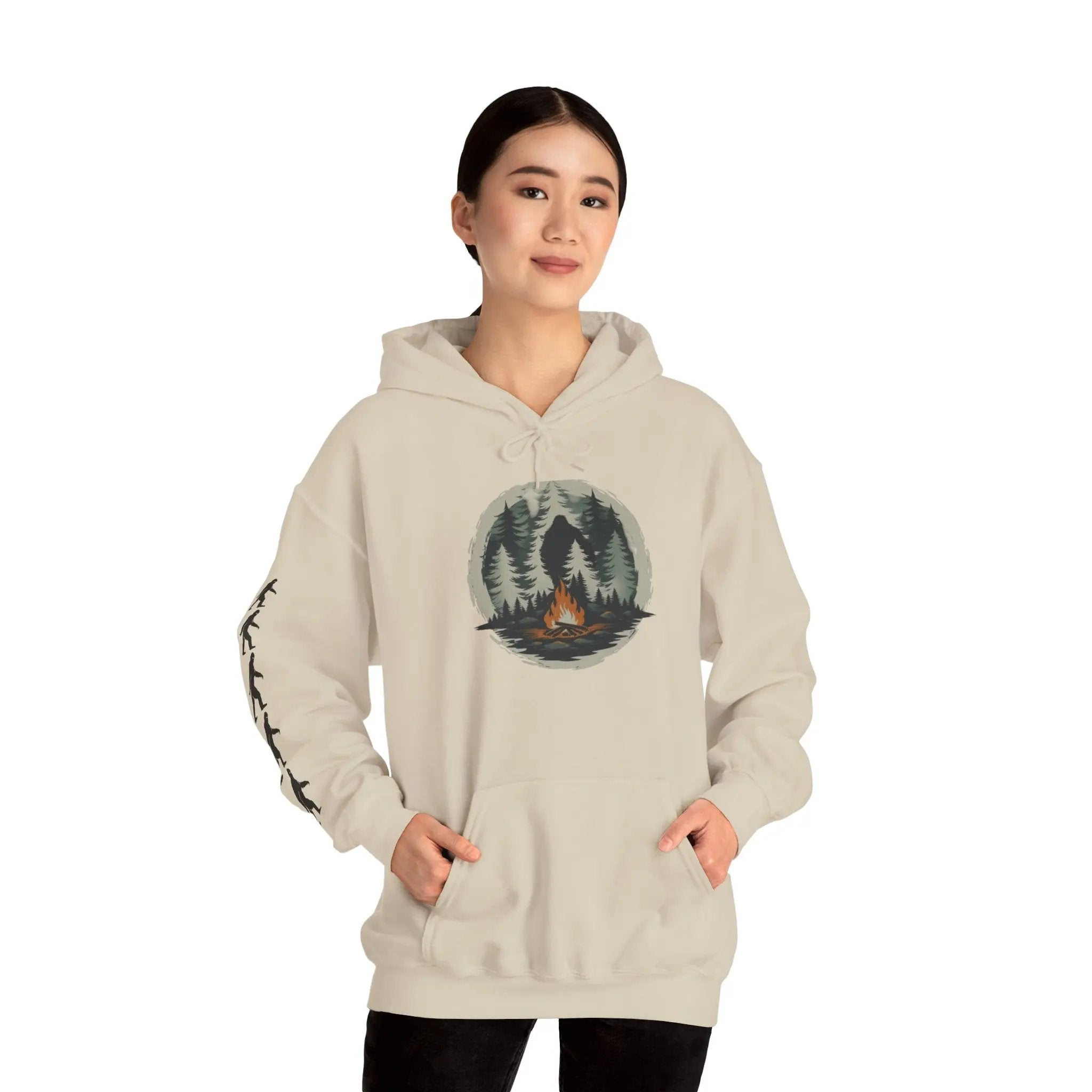 Person wearing a beige hoodie with a forest and campfire graphic and black bigfoot silhouettes on the right sleeve on a white background on a white background