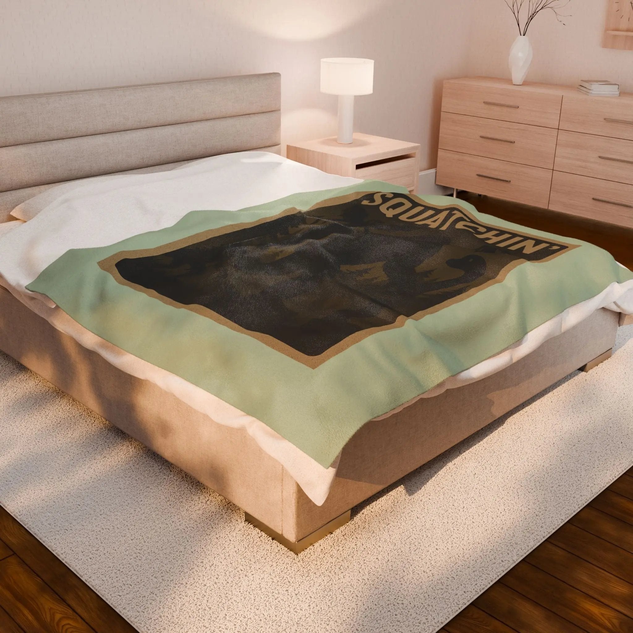 Light green fleece blanket with 'Squatchin' text across the top with silhouettes of bigfoot, loch Ness, werewolf, mothman, and an alien on a bed in a bedroom setting.
