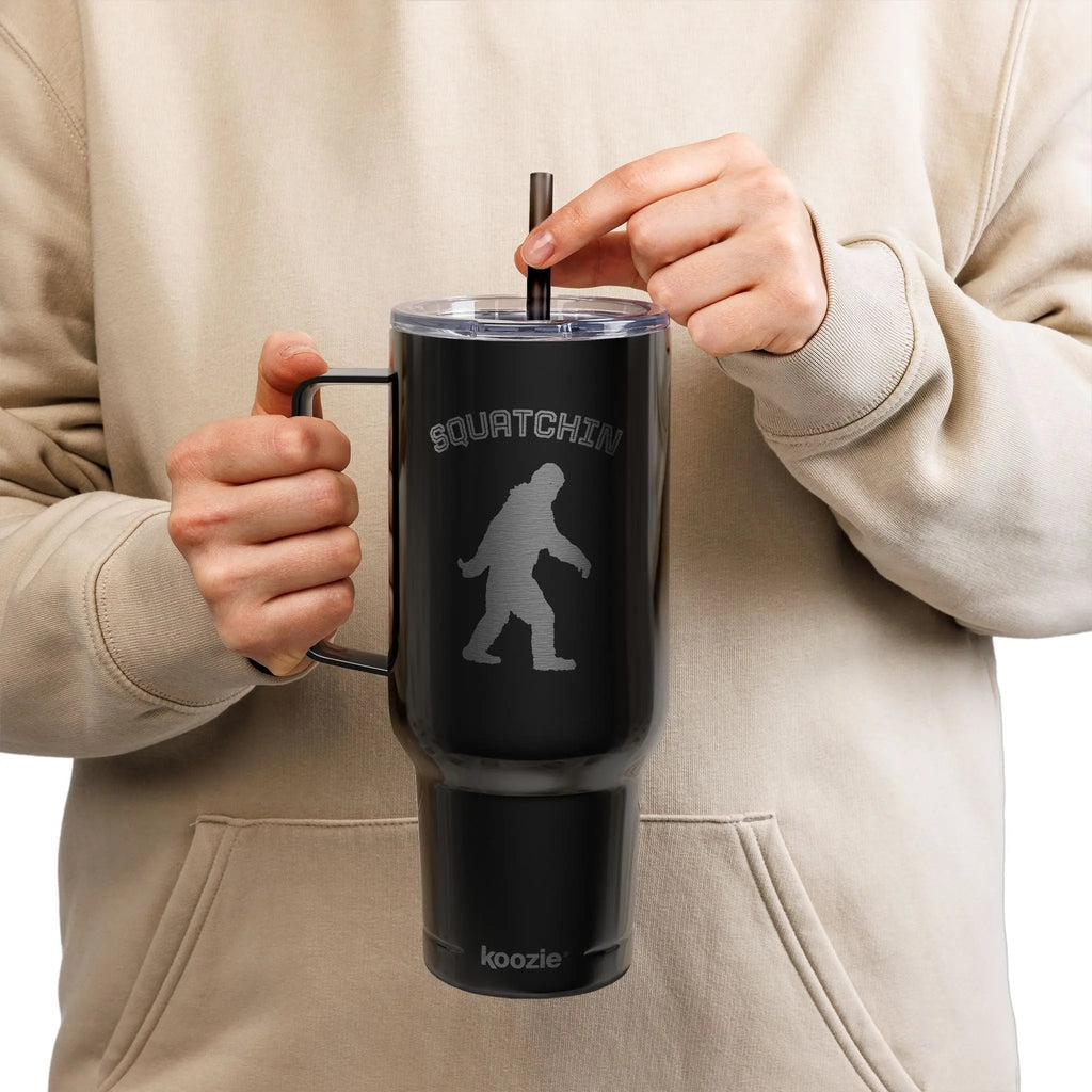 Person holding a black tumbler with 'Squatchin' and a silhouette of Bigfoot on a beige background