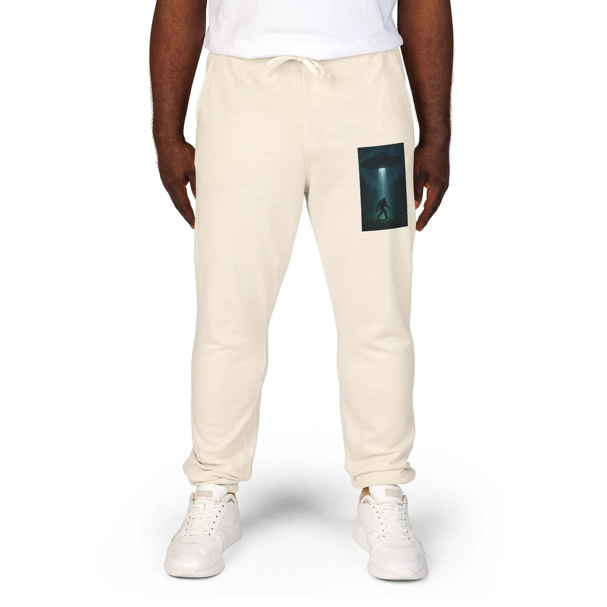 Beige sweatpants with a small graphic of a UFO with a beam of light on a bigfoot silhouette on the left leg on a white background