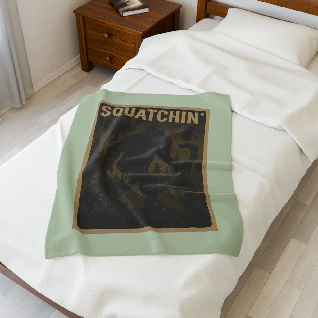 Light green fleece blanket with 'Squatchin' text across the top with silhouettes of bigfoot, loch Ness, werewolf, mothman, and an alien on a bed in a bedroom setting.