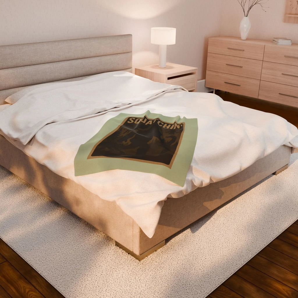 Light green fleece blanket with 'Squatchin' text across the top with silhouettes of bigfoot, loch Ness, werewolf, mothman, and an alien on a bed in a bedroom setting.