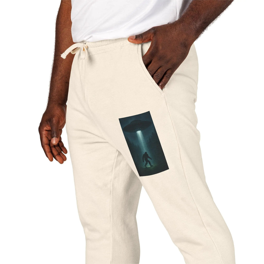 Beige sweatpants with a small graphic of a UFO with a beam of light on a bigfoot silhouette on the left leg worn by a person.