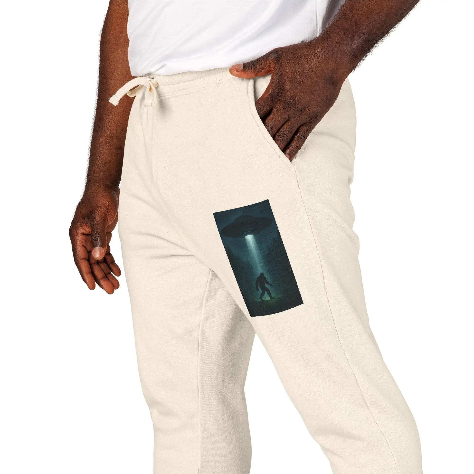Beige sweatpants with a small graphic of a UFO with a beam of light on a bigfoot silhouette on the left leg worn by a person.